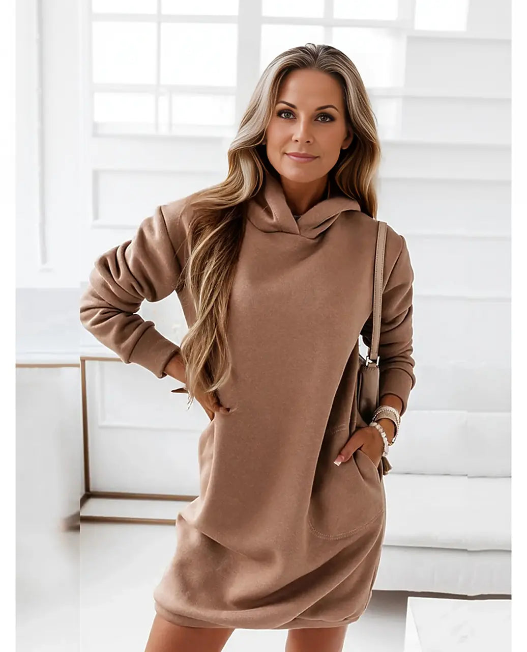 Frau in braunem Hoodie-Kleid posiert in modernem, hellem Raum. Modetrend, lässiger Stil, Damenmode, Herbstoutfit, bequemes Kleid.