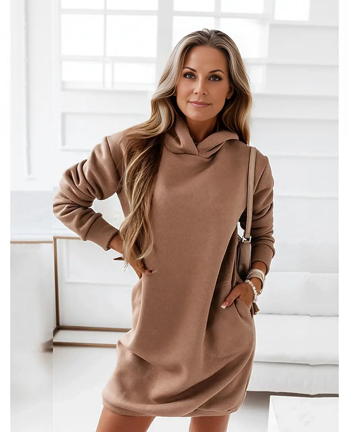 Frau in braunem Hoodie-Kleid posiert in modernem, hellem Raum. Modetrend, lässiger Stil, Damenmode, Herbstoutfit, bequemes Kleid.