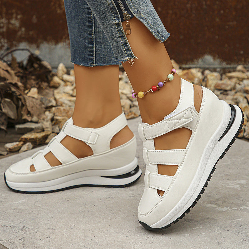 Weiße Plateau-Sandalen mit Klettverschluss an Frauenfüßen, kombiniert mit Jeans und buntem Fußkettchen. Modische Sommerschuhe für Damen.