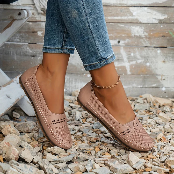 Damenfüße in rosa Mokassins auf Kieselsteinen, Jeans hochgekrempelt, rustikaler Holzhintergrund. Bequeme, stilvolle Freizeitschuhe für den Alltag.