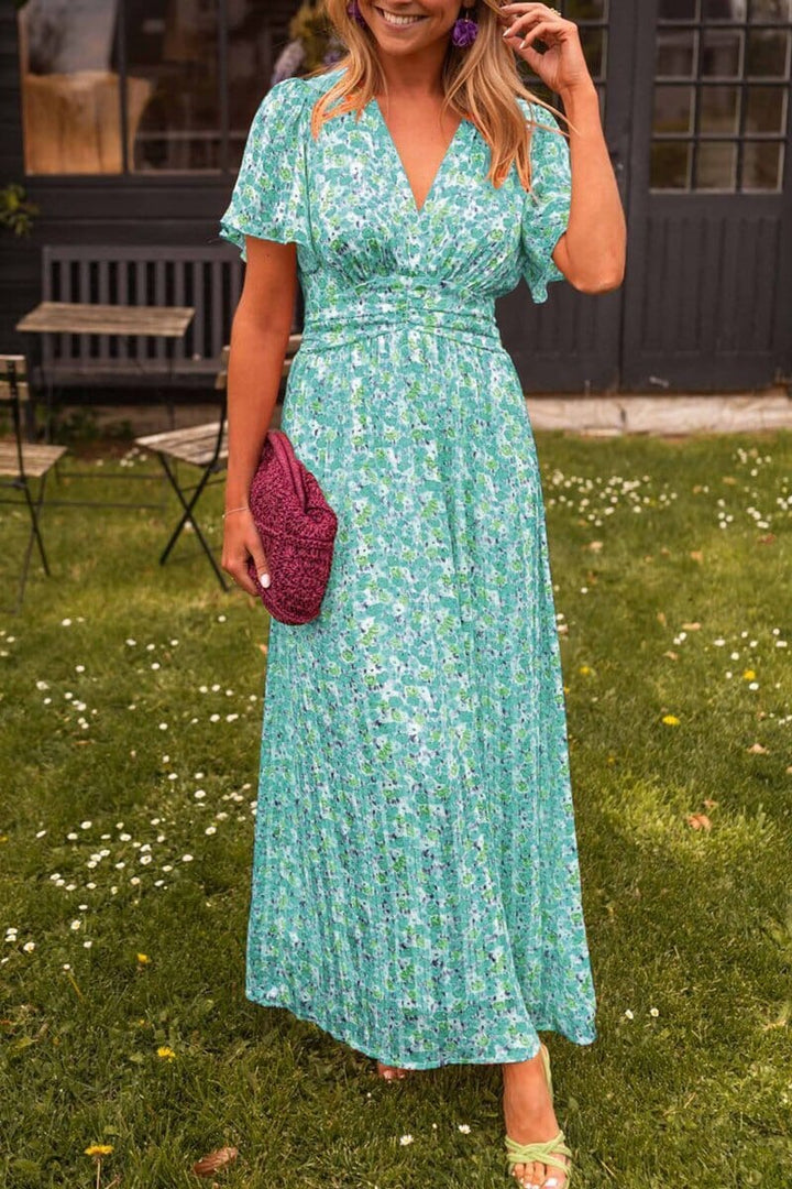 Frau in langem, geblümtem Sommerkleid in Grün und Blau, hält eine lila Clutch, steht auf einer Wiese vor einem dunklen Gebäude.