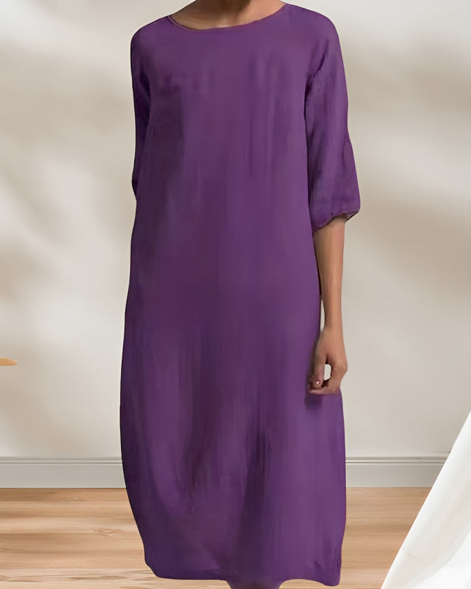 Frau in lila Kleid, knielang, mit halblangen Ärmeln, steht auf Holzboden vor neutralem Hintergrund. Elegantes, minimalistisches Design.