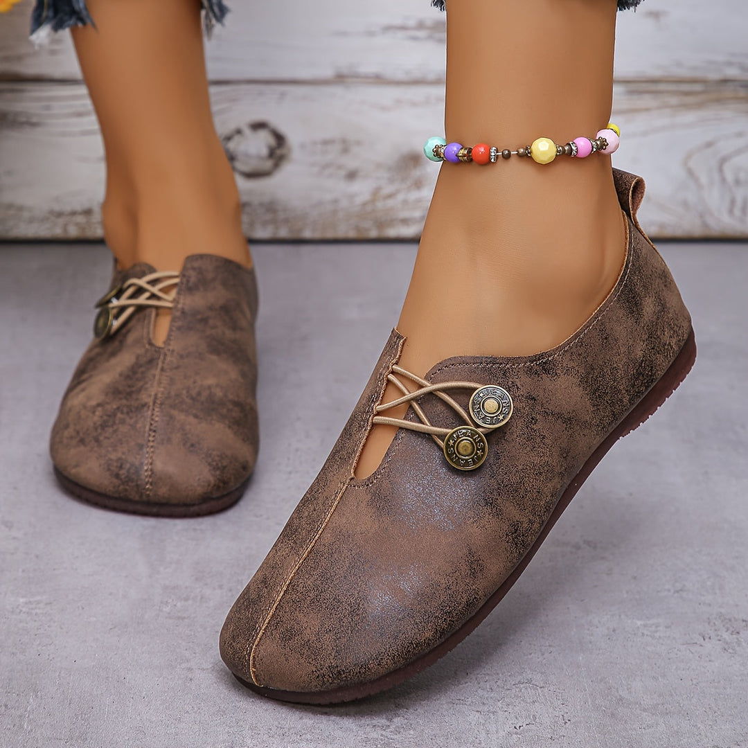 Braune Damen-Lederslipper mit dekorativen Knöpfen und bunter Perlenkette am Knöchel, modische Freizeitschuhe auf grauem Boden.