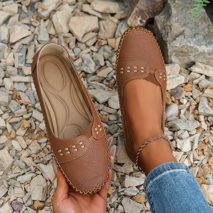 Braune Damen-Loafer aus Leder mit Perforationen, getragen auf Kieselsteinen. Bequeme, stilvolle Schuhe für Freizeit und Alltag.