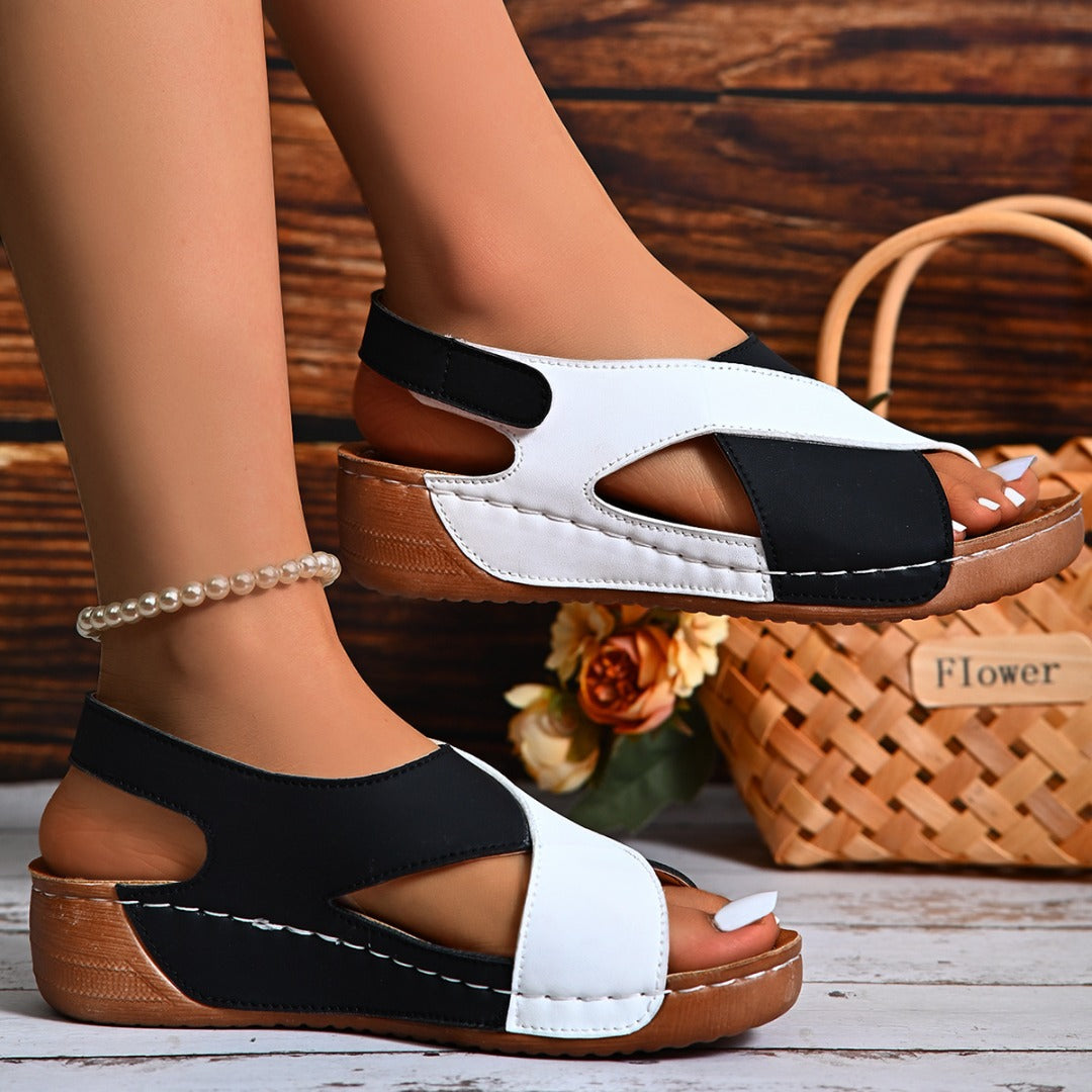 Schwarze und weiße Sandalen mit Keilabsatz, getragen von einer Person mit Perlenarmband, vor einem Korb mit Blumen auf Holzboden.