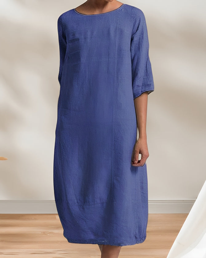 Frau in einem blauen Leinenkleid, knielang, mit 3/4-Ärmeln, steht auf einem Holzboden. Elegantes, minimalistisches Design für Sommermode.