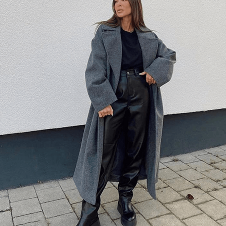Frau in grauem Wollmantel, schwarzem Pullover und Lederhose, steht auf gepflastertem Boden vor weißer Wand. Modetrend Herbst/Winter.