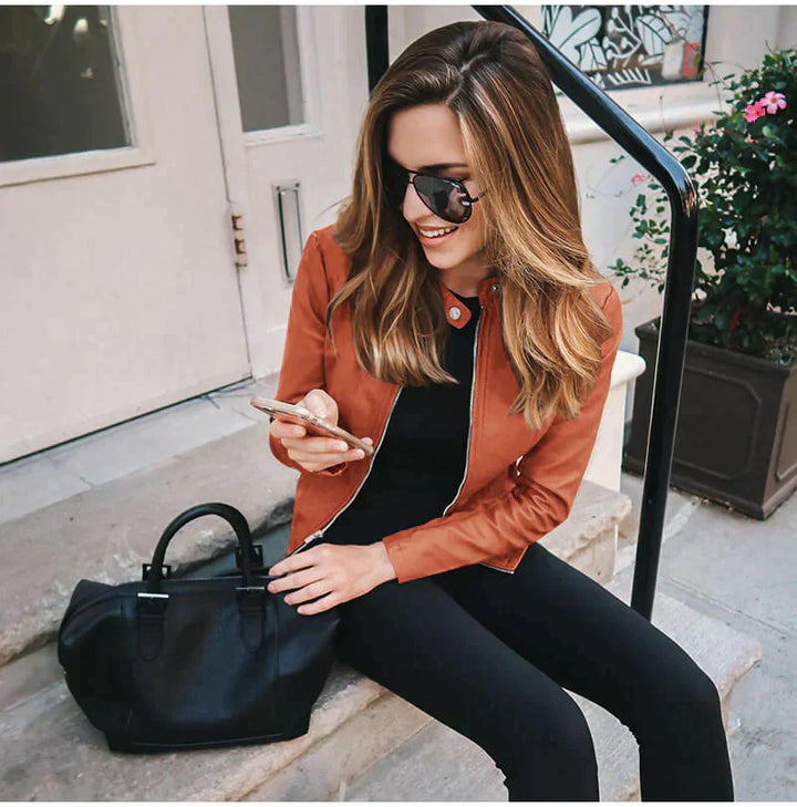 Frau mit Sonnenbrille und brauner Jacke sitzt auf Treppe, hält Smartphone. Schwarze Handtasche neben ihr. Urbaner Stil, Mode, Lifestyle.