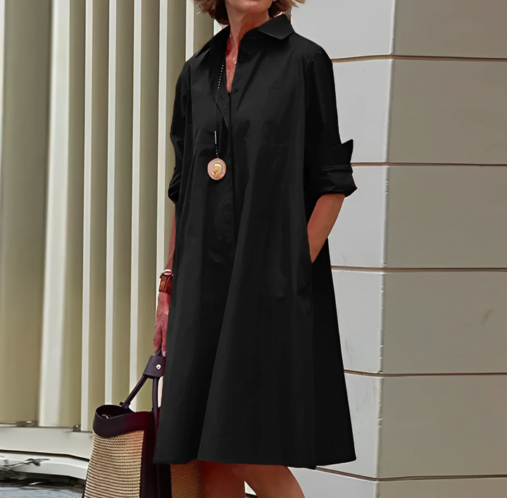 Eine Frau in einem eleganten, schwarzen Hemdkleid mit langen Ärmeln, trägt eine Halskette und hält eine Tasche. Urbaner Hintergrund mit moderner Architektur.