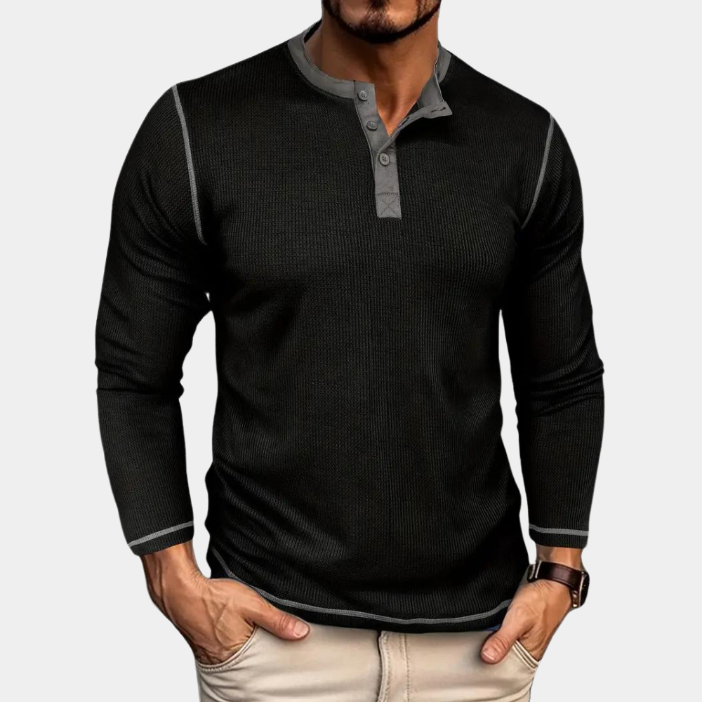 Mann trägt schwarzes Langarm-Henley-Shirt mit grauen Akzenten, beige Hose. Modischer Herrenstil, Freizeitkleidung, Herbstmode.