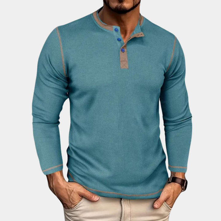 Mann trägt blaues Langarm-Henley-Shirt mit Knopfleiste, kombiniert mit beigen Hosen. Modischer Freizeitlook für Herrenmode.