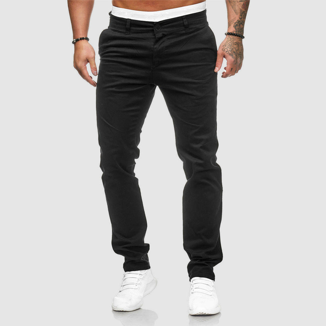 Mann in schwarzer Chinohose und weißen Sneakers vor grauem Hintergrund. Modische Herrenbekleidung, lässiger Stil, moderne Herrenmode.