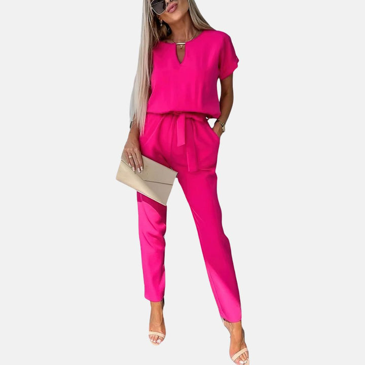 Frau in pinkem Jumpsuit mit Gürtel, hält eine beige Clutch, trägt Sonnenbrille und Sandalen. Modetrend, Damenmode, eleganter Look.