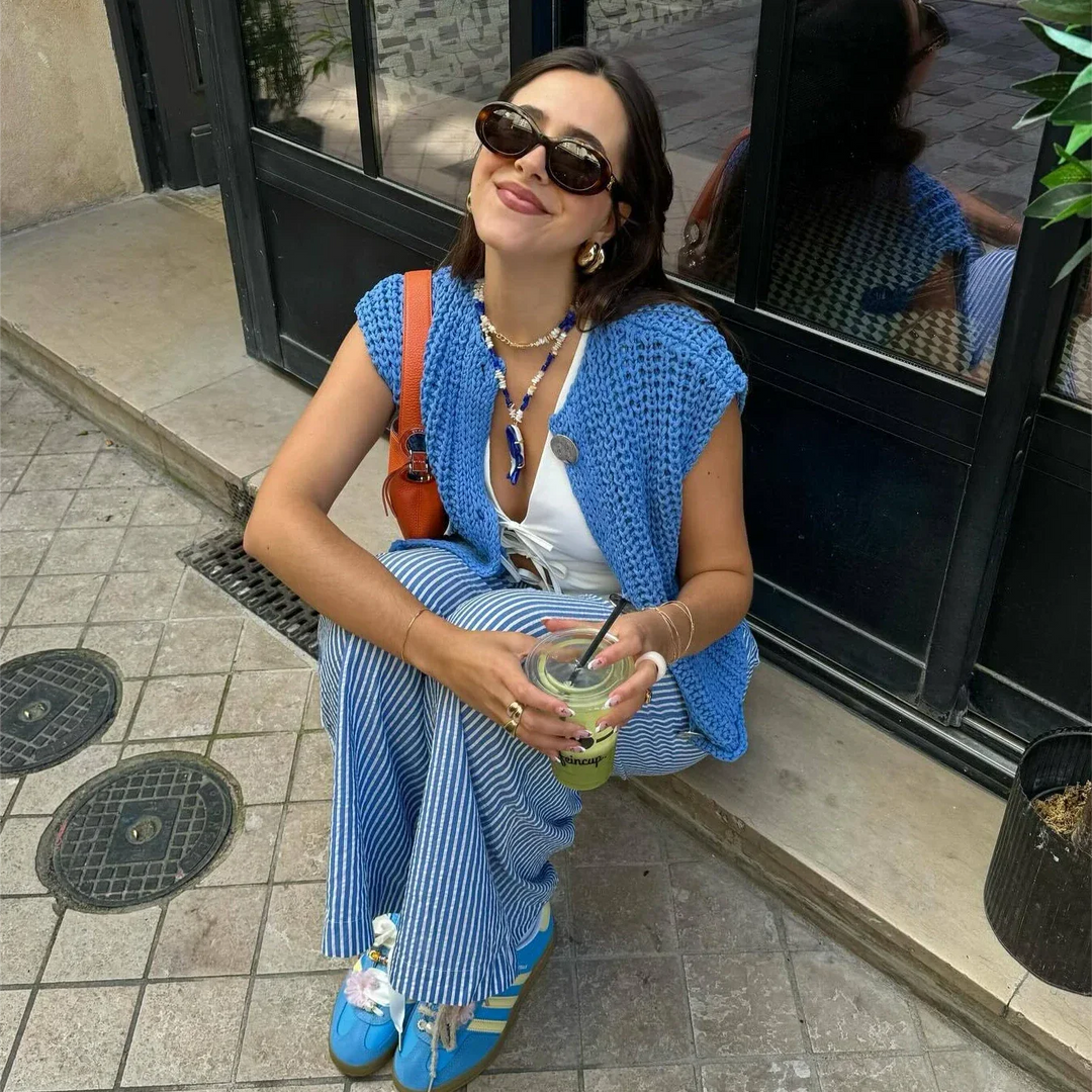 Frau in blauer Strickweste und gestreifter Hose sitzt lächelnd mit Sonnenbrille und Getränk auf dem Bürgersteig. Modetrend, lässiger Sommerlook.