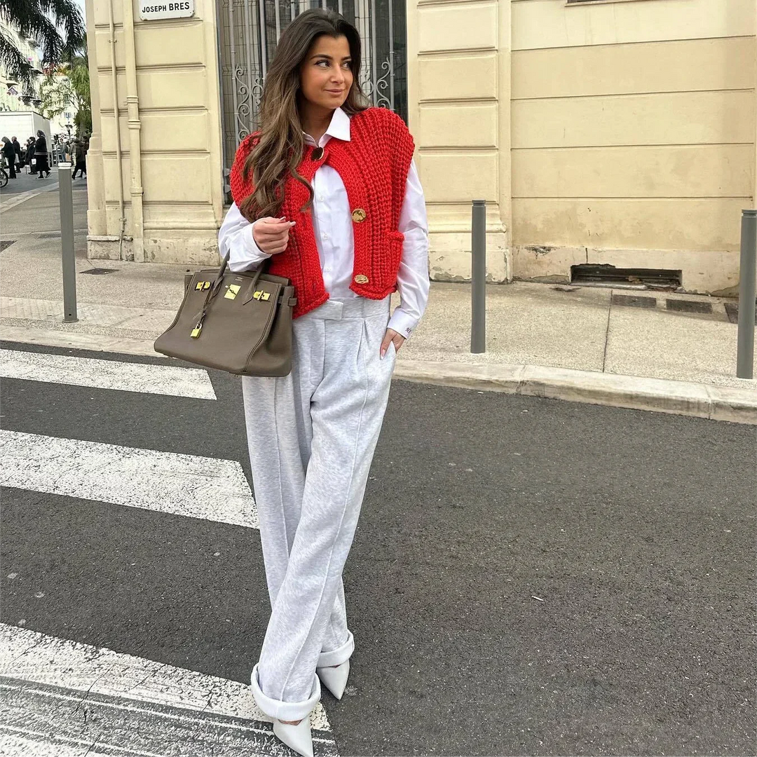 Frau in weißem Hemd und grauer Hose mit roter Strickweste und großer brauner Handtasche steht auf Gehweg. Straßenmode, Herbstoutfit, urbaner Stil.