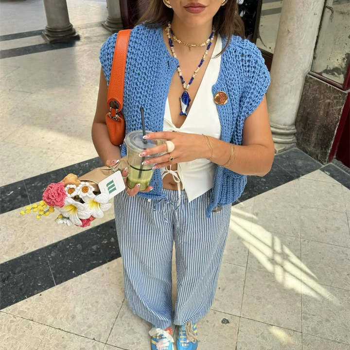 Frau in gestreifter Hose und blauem Strickoberteil hält Eiskaffee und Blumenstrauß. Modetrend, Sommeroutfit, lässiger Stil.