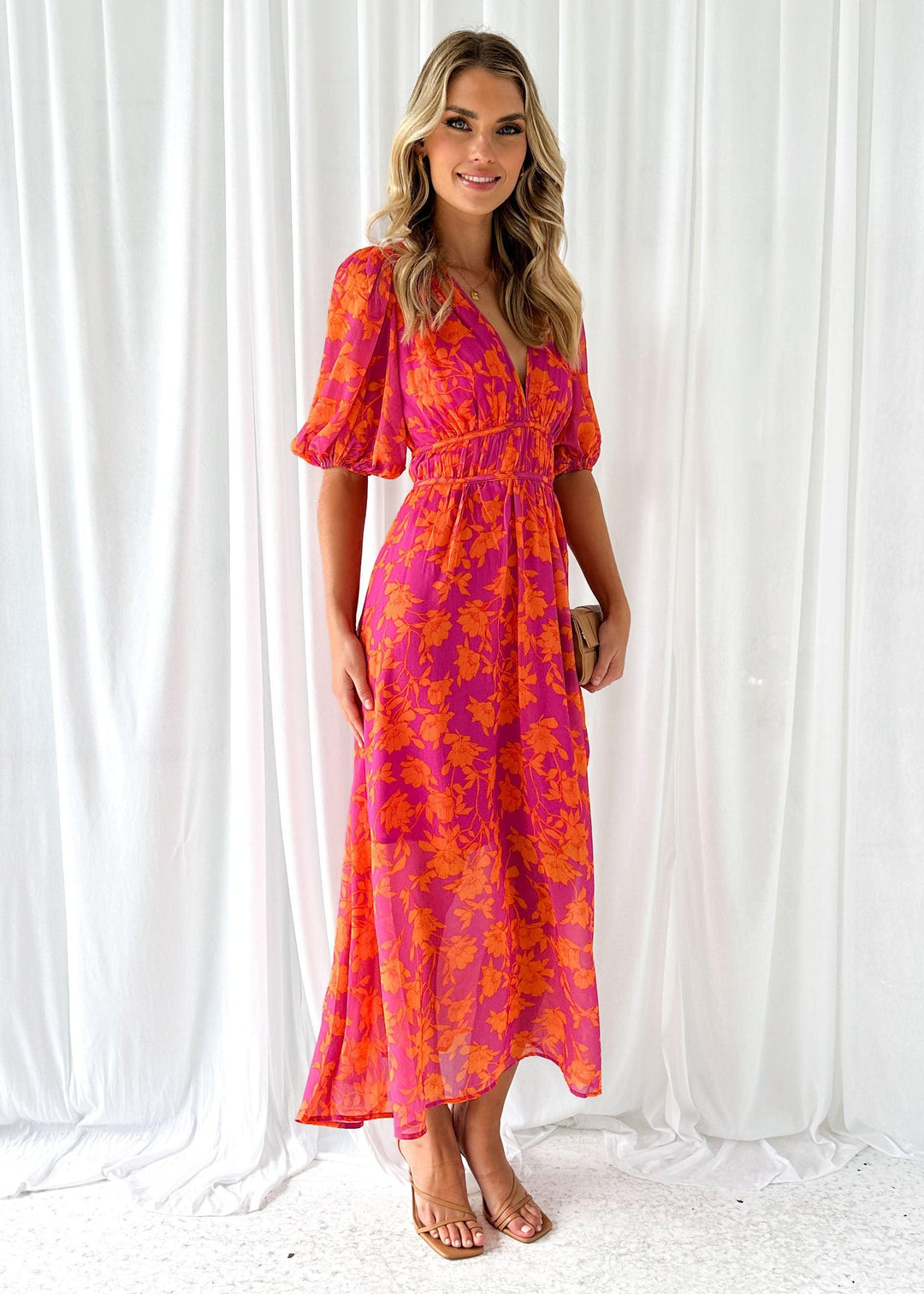 Frau in langem, pink-orange geblümtem Kleid vor weißem Hintergrund, lächelt. Sommermode, elegantes Maxikleid, florales Design, Damenbekleidung.