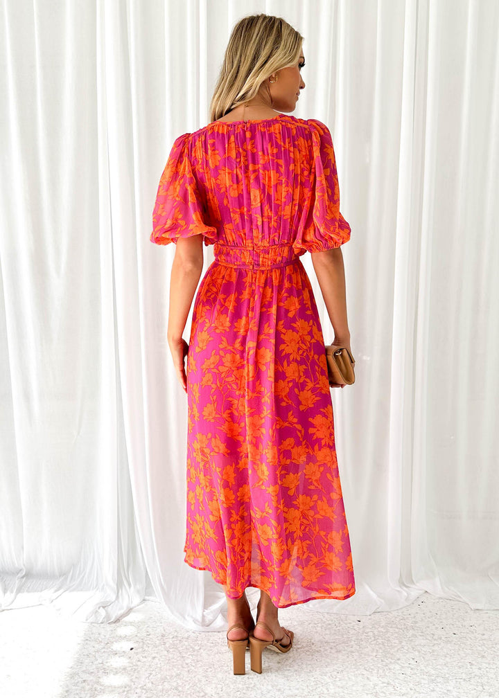 Frau in langem, pink-orange geblümtem Kleid mit Puffärmeln, steht vor weißen Vorhängen. Sommermode, elegantes Design, Rückansicht.