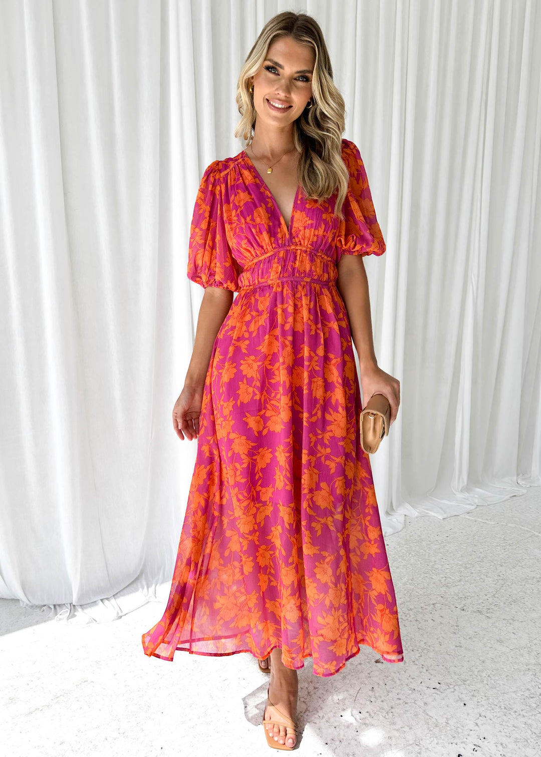 Frau in einem langen, floralen Sommerkleid in Pink und Orange, lächelnd vor weißen Vorhängen. Elegantes Outfit, modisches Design, Sommermode.