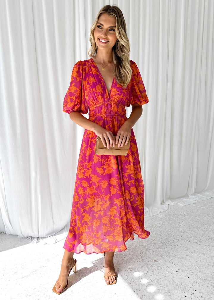 Frau in einem pink-orange geblümten Kleid, lächelnd, mit Clutch in der Hand, vor weißem Vorhang. Sommermode, elegantes Damenoutfit, florales Design.