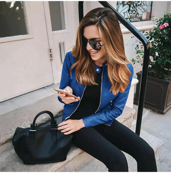 Frau mit blauer Jacke und Sonnenbrille sitzt auf Treppe, schaut auf Smartphone, schwarze Tasche neben ihr. Urbaner Stil, Mode, Outdoor.
