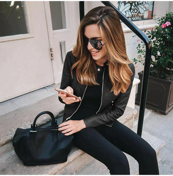 Frau mit Sonnenbrille und Lederjacke sitzt auf Treppe, hält Smartphone. Schwarze Handtasche neben ihr. Urbaner Stil, Mode, Lifestyle.