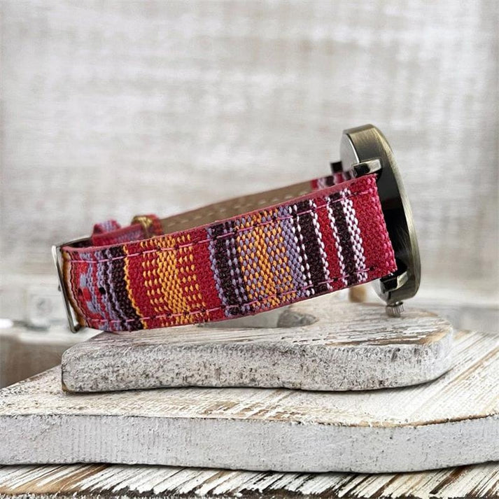 Bunte Armbanduhr mit gewebtem Stoffarmband in Rot- und Lilatönen, auf rustikalem Holzuntergrund. Handgefertigte Uhr, modisches Accessoire.