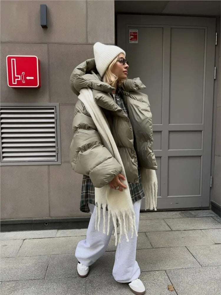 Frau in beiger Daunenjacke, weißer Mütze und Schal, grauer Jogginghose und Sonnenbrille vor grauer Tür. Wintermode, Streetstyle, urbaner Look.