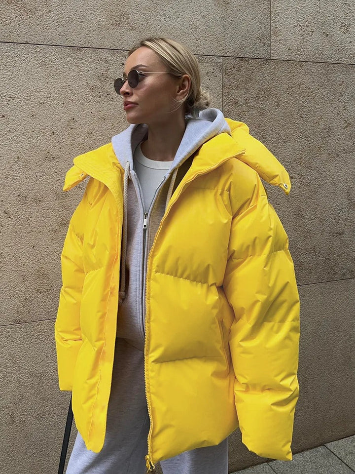 Frau in gelber Daunenjacke, grauem Hoodie und Sonnenbrille vor beiger Wand. Wintermode, Streetstyle, trendige Outfits, warme Kleidung.