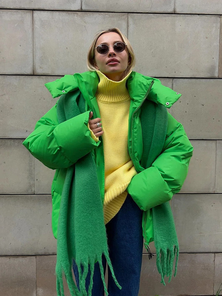Frau in grüner Daunenjacke und gelbem Rollkragenpullover vor Betonwand, trägt Sonnenbrille und grünen Schal. Wintermode, Streetstyle, trendig.