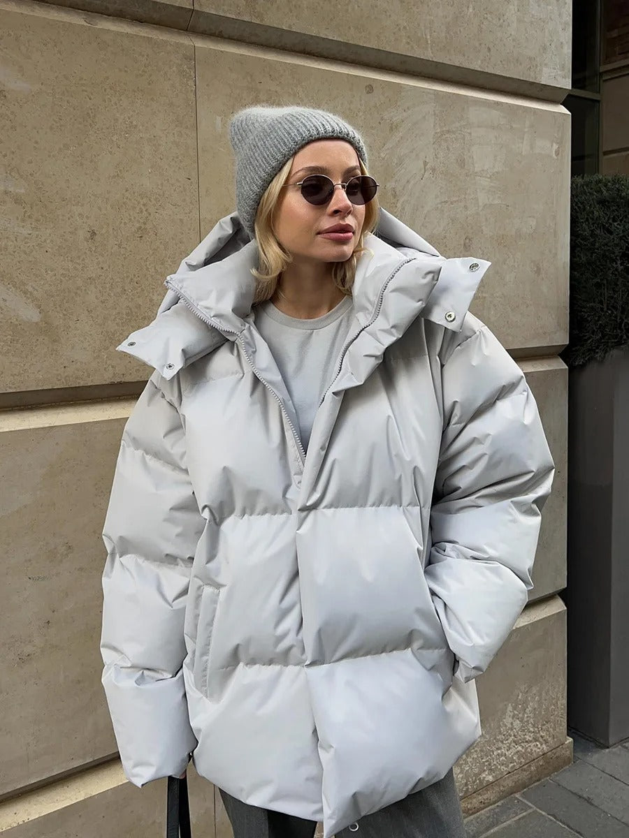 Frau in grauer Daunenjacke und Mütze, trägt Sonnenbrille, steht vor beiger Wand. Wintermode, urbane Kleidung, stilvoller Look.