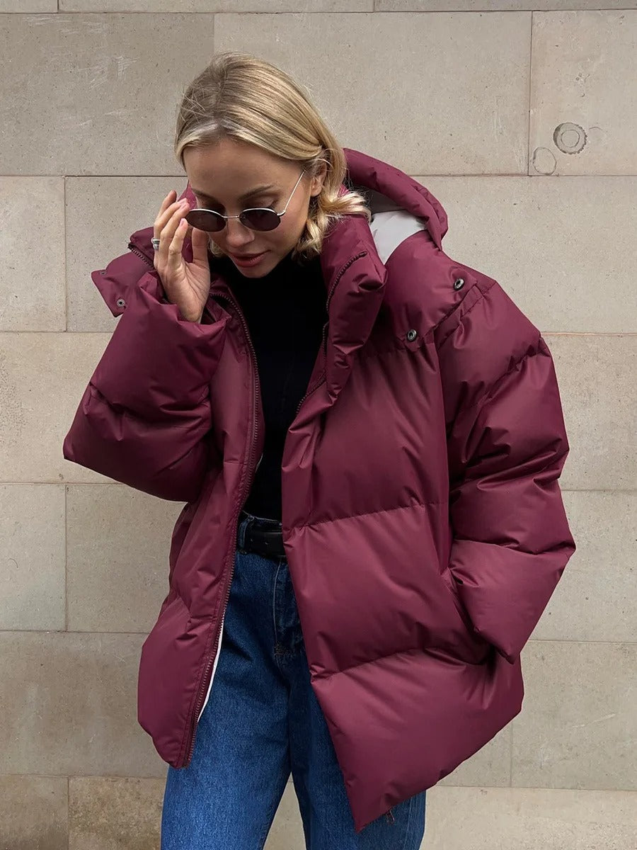 Frau in burgunderroter Daunenjacke, Sonnenbrille, Jeans vor beiger Wand. Wintermode, stylische Damenbekleidung, warme Jacke.