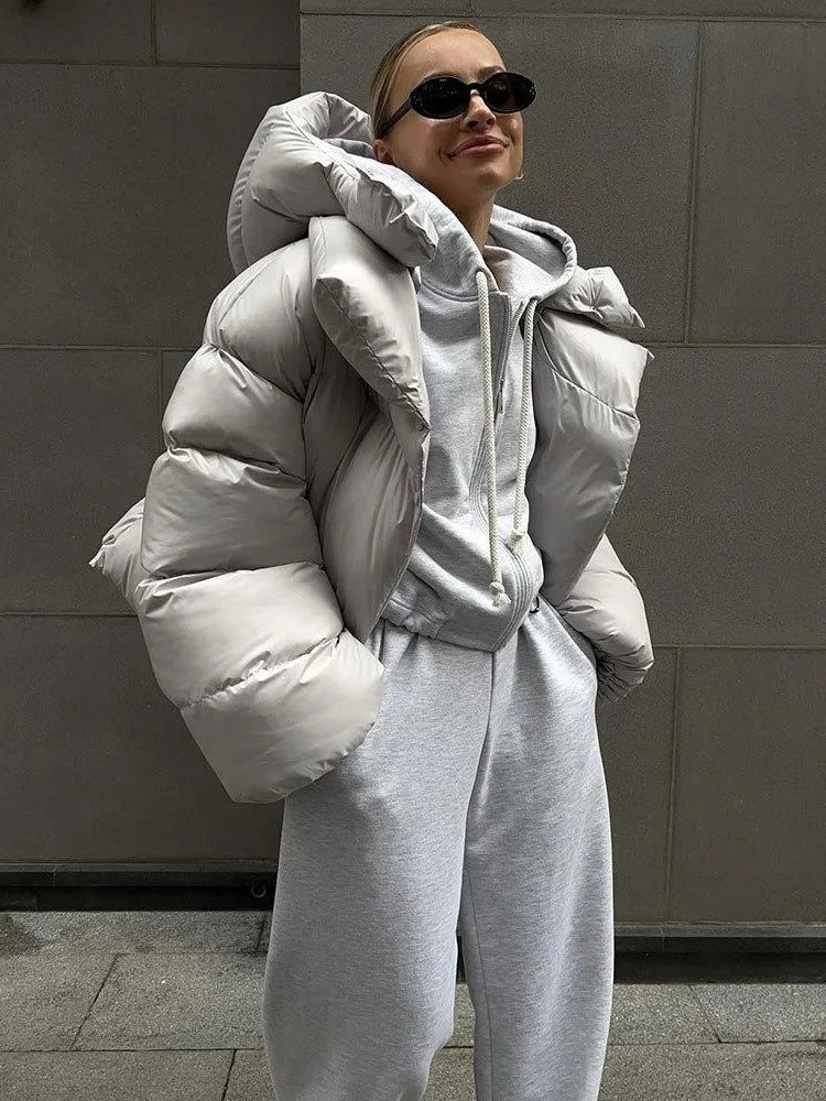 Frau in grauem Hoodie und Jogginghose mit weißer Daunenjacke und Sonnenbrille, lässiger Streetstyle, moderne Wintermode, urbaner Hintergrund.