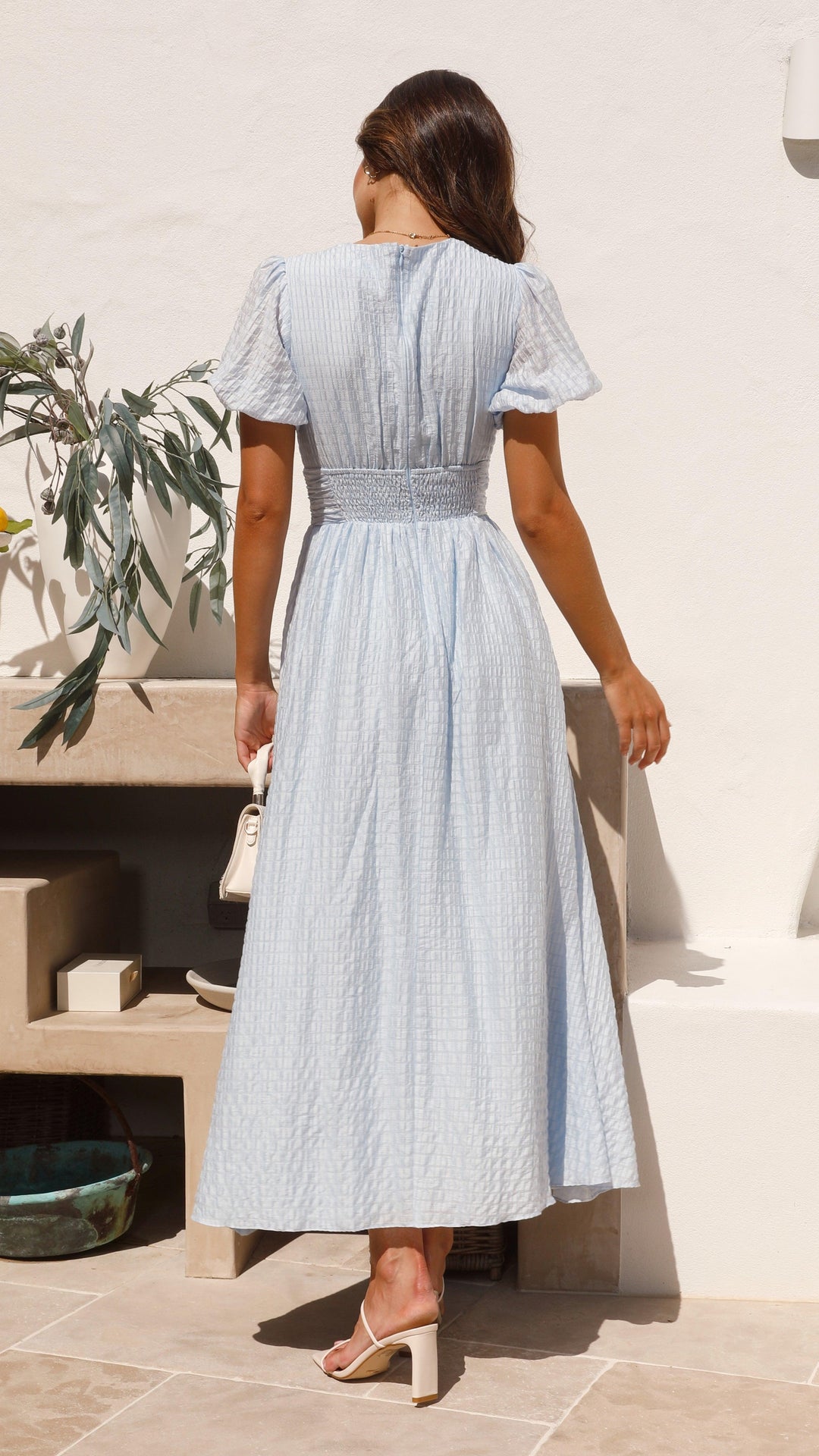 Frau in hellblauem Sommerkleid mit Puffärmeln, steht auf Terrasse. Elegantes, luftiges Design, ideal für warme Tage. Mode, Damenbekleidung, Sommerstil.