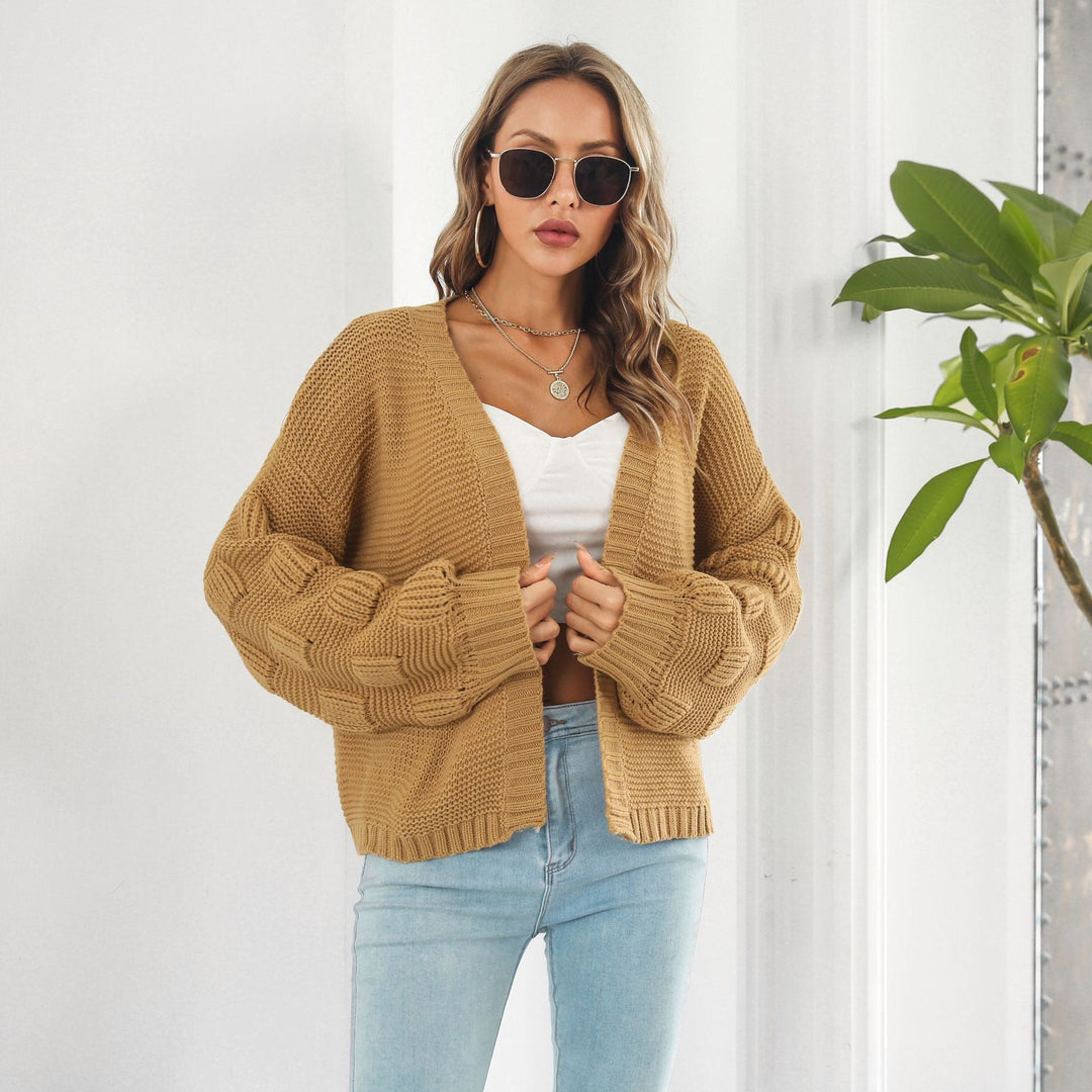 Frau mit Sonnenbrille trägt beige Strickjacke und weißes Top, steht vor weißer Wand mit Pflanze. Modetrend, Damenmode, Strickjacke, lässiger Look.