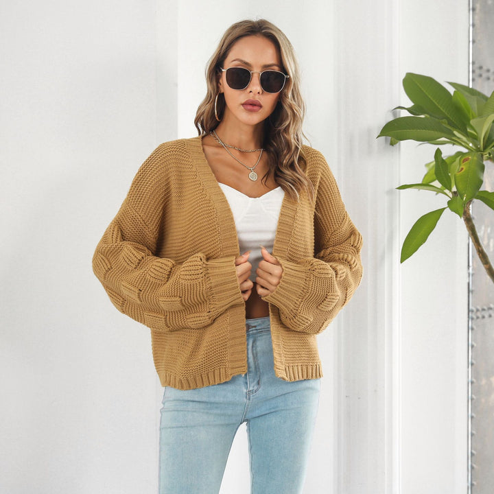 Frau mit Sonnenbrille trägt beige Strickjacke und weißes Top, steht vor weißer Wand mit Pflanze. Modetrend, Damenmode, Strickjacke, lässiger Look.