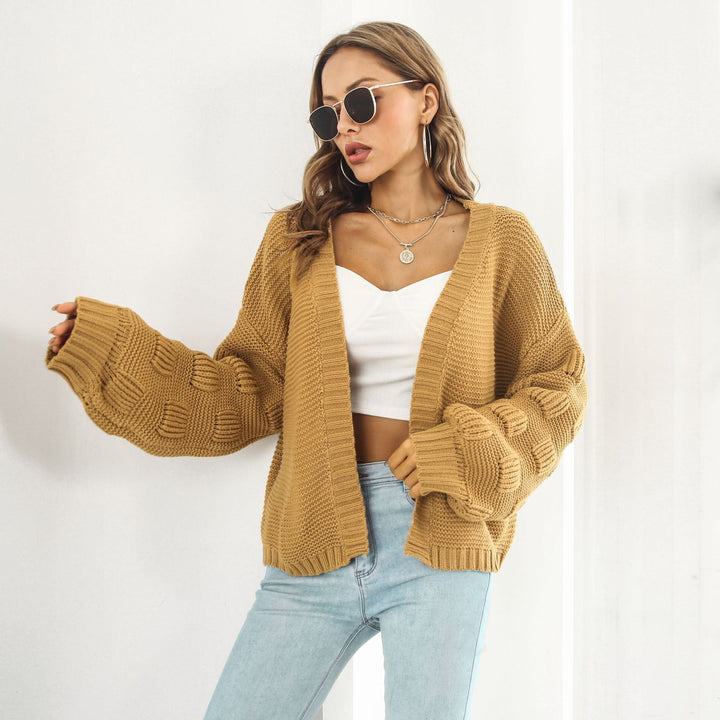 Frau in modischer, beiger Strickjacke mit Zopfmuster, weißem Top und blauer Jeans, trägt Sonnenbrille und Schmuck, steht vor hellem Hintergrund.