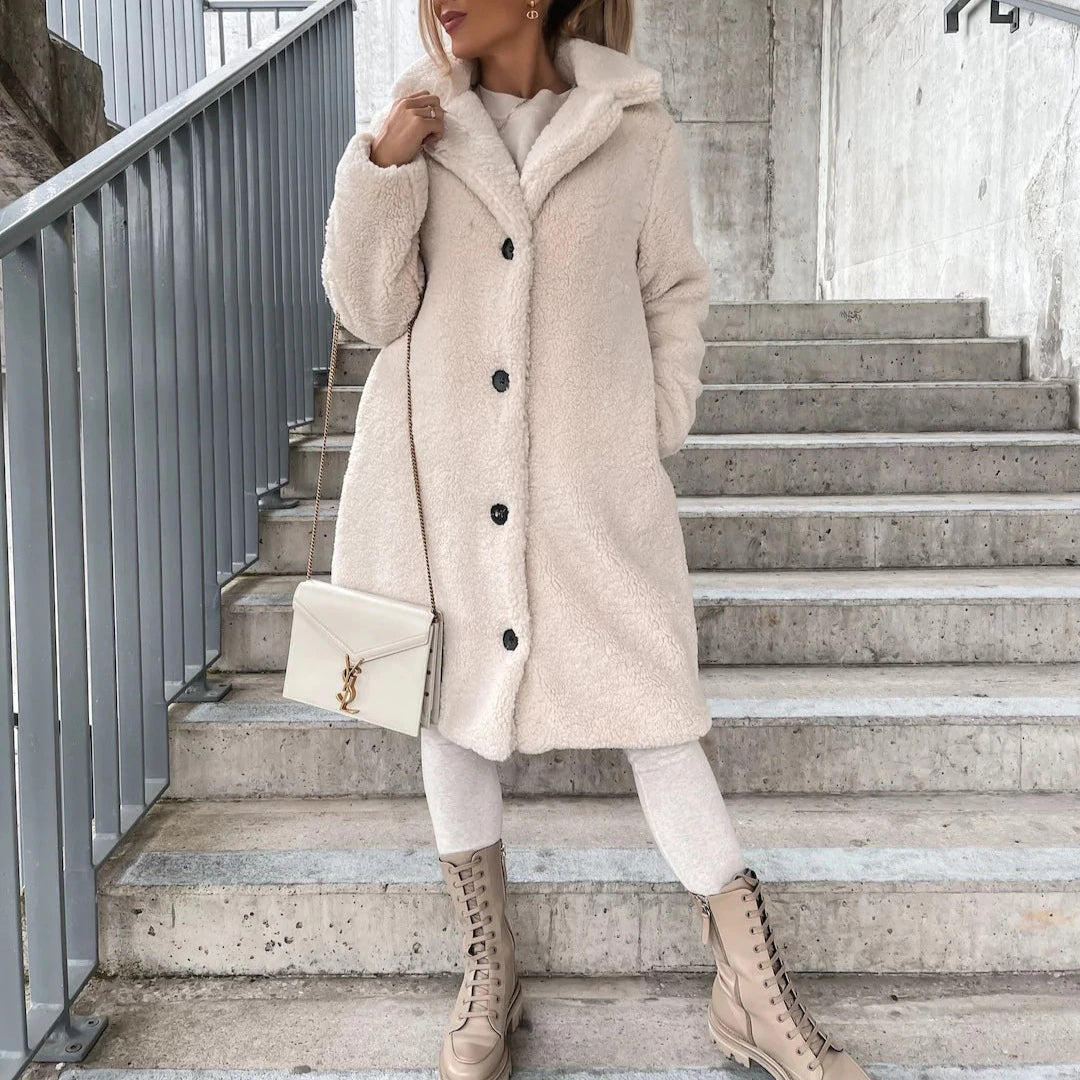 Frau in beigem Teddy-Mantel auf Treppe, trägt cremefarbene Stiefel und hält eine Designer-Handtasche. Wintermode, eleganter Look, urbane Kulisse.