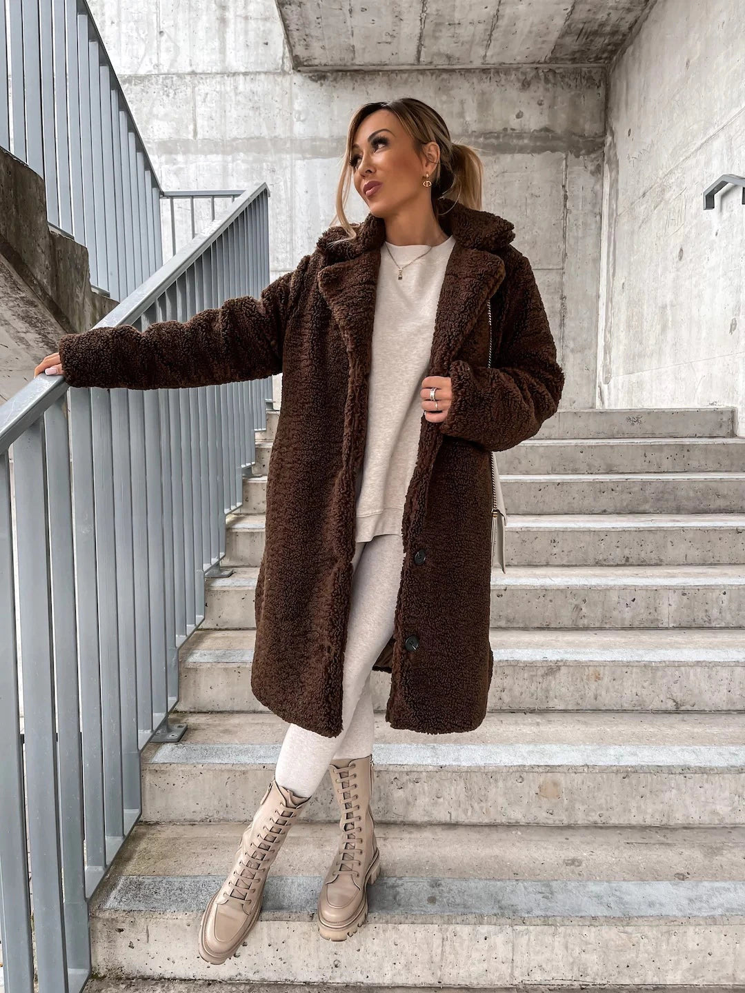 Frau in braunem Teddy-Mantel und beiger Kleidung steht auf Treppe. Modetrend, Wintermode, urbaner Stil, gemütlicher Look, Streetwear.