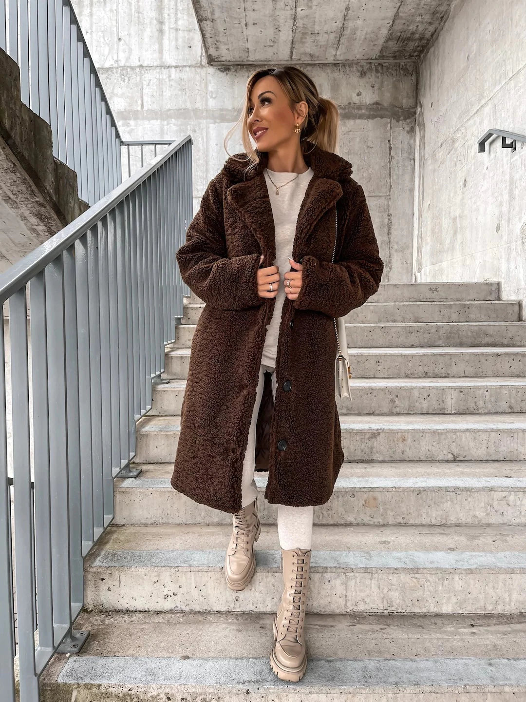Frau in braunem Teddy-Mantel auf Treppe, beige Stiefel, urbaner Hintergrund. Wintermode, stilvoll, modern, Outdoor-Look.