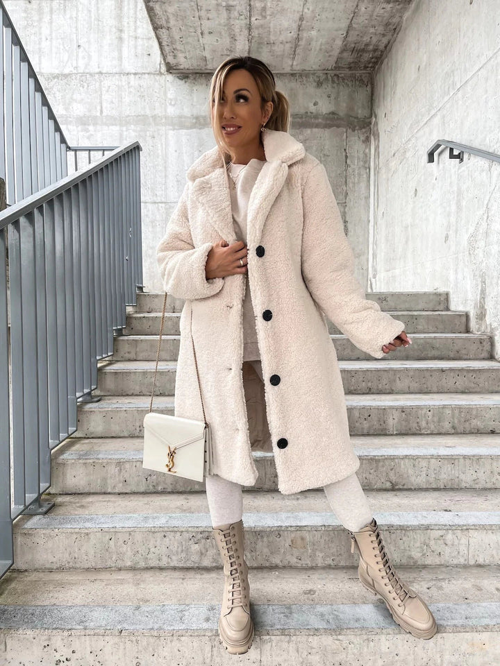 Frau in weißem Teddy-Mantel auf Treppe, trägt beige Stiefel und weiße Tasche. Wintermode, eleganter Look, urbane Kulisse.