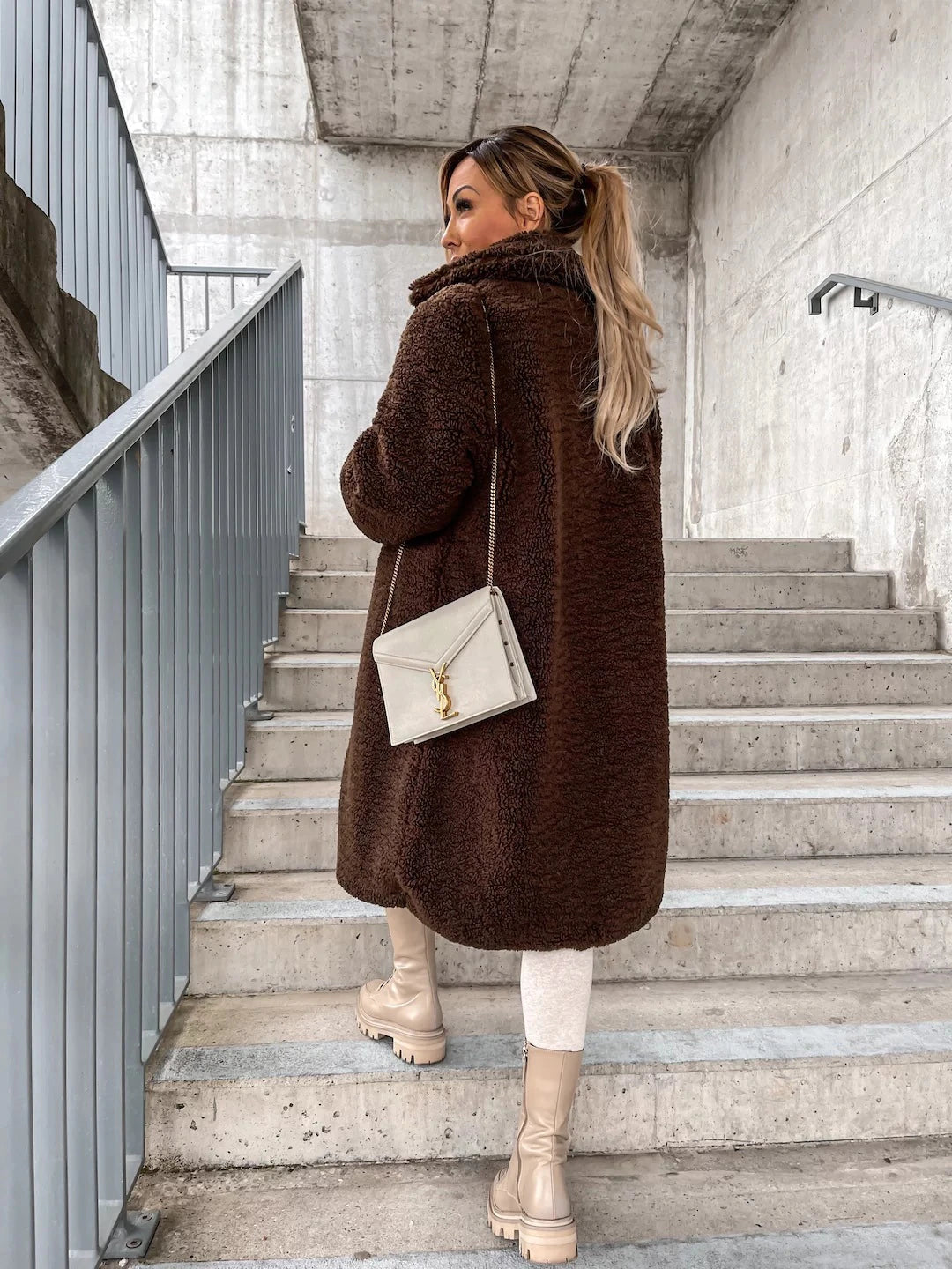 Frau in braunem Teddy-Mantel auf Treppe, beige Stiefel, cremefarbene Umhängetasche. Wintermode, urbane Kulisse, stilvoller Look.
