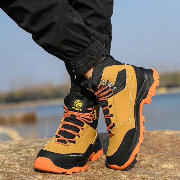 Braune Wanderschuhe mit orangefarbenen Sohlen und Schnürsenkeln auf Felsen, getragen von Person in schwarzer Hose. Outdoor, Trekking, robustes Design.