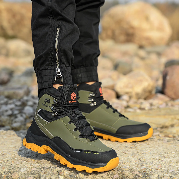 Grüne Outdoor-Wanderschuhe mit orangefarbener Sohle auf steinigem Untergrund, getragen von Person in schwarzer Hose. Robuste Trekkingstiefel für Herren.