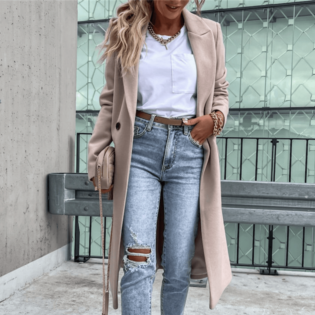 Frau in beigem Mantel, weißem Shirt und zerrissenen Jeans posiert vor modernem Gebäude. Modetrend, Streetstyle, Herbstmode, lässiger Look.