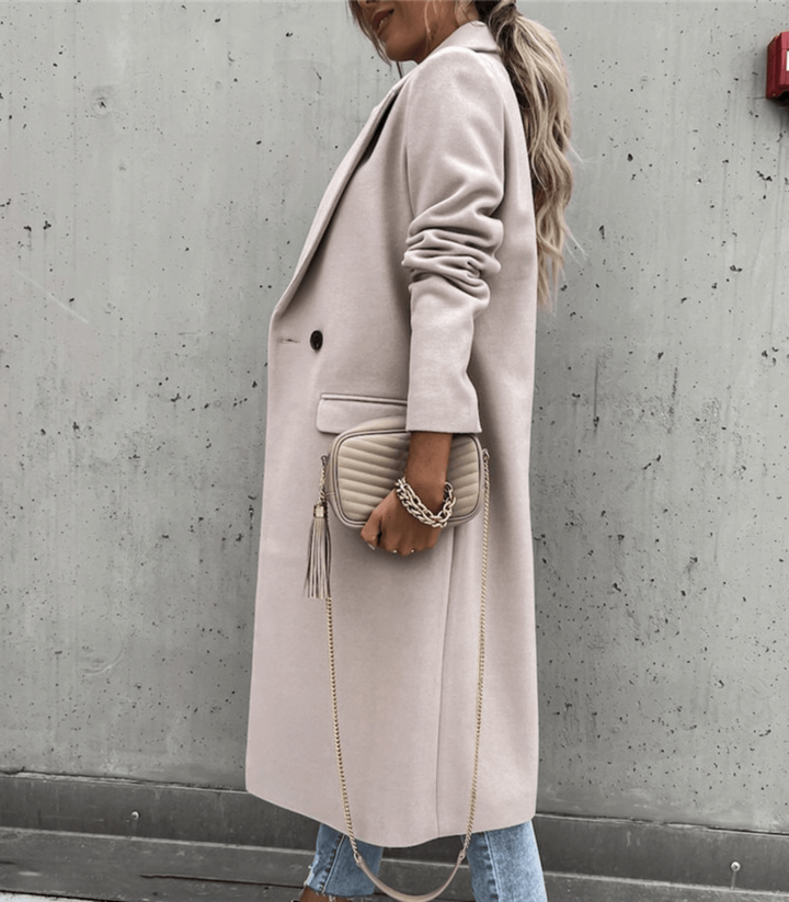 Frau in beigem Mantel mit gesteppter Handtasche und goldener Kette, steht vor grauer Betonwand. Modetrend, Herbstmode, eleganter Look.