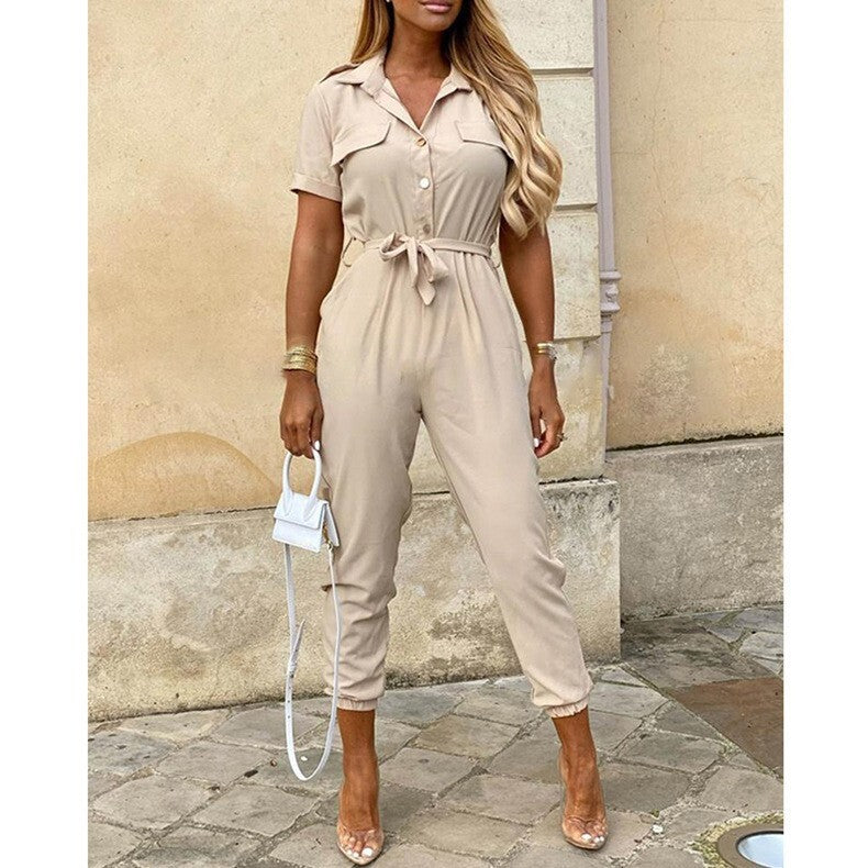 Frau in beigem Jumpsuit mit Gürtel, hält weiße Handtasche. Modetrend, eleganter Einteiler, Sommeroutfit, stilvolle Damenmode, urbane Kulisse.