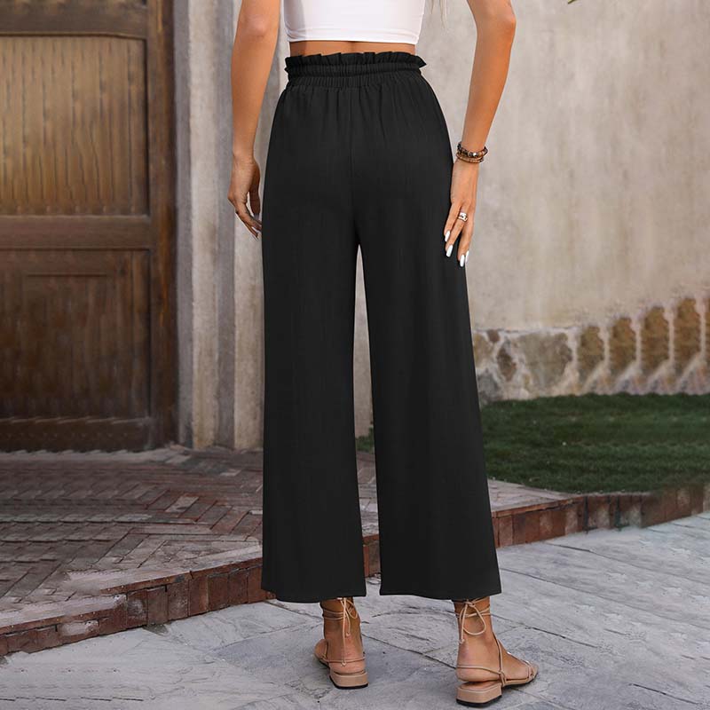 Frau in schwarzer, hoch taillierter, weiter Hose und weißem Top, steht draußen vor Holztür. Sommermode, lässiger Stil, elegante Damenbekleidung.