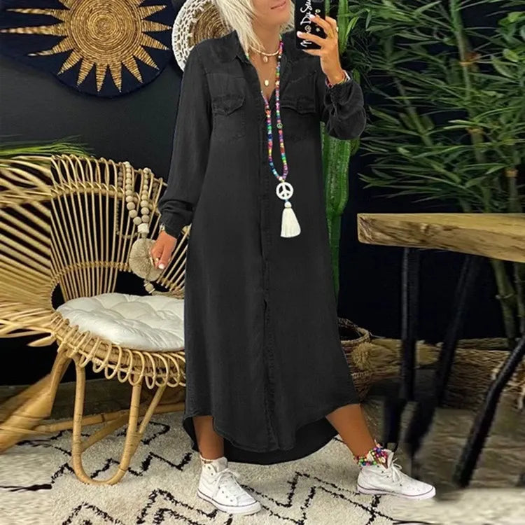 Frau in schwarzem Maxikleid mit langen Ärmeln, bunter Halskette und weißen Sneakers, steht in modernem Wohnzimmer mit Rattanmöbeln und Pflanzen.