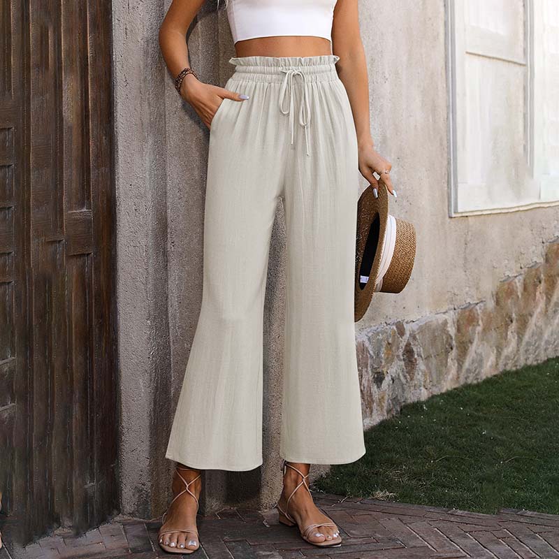 Frau in beiger Leinenhose und weißem Crop-Top, hält Strohhut. Sommermode, lässiger Stil, bequeme Kleidung, trendige Damenbekleidung.